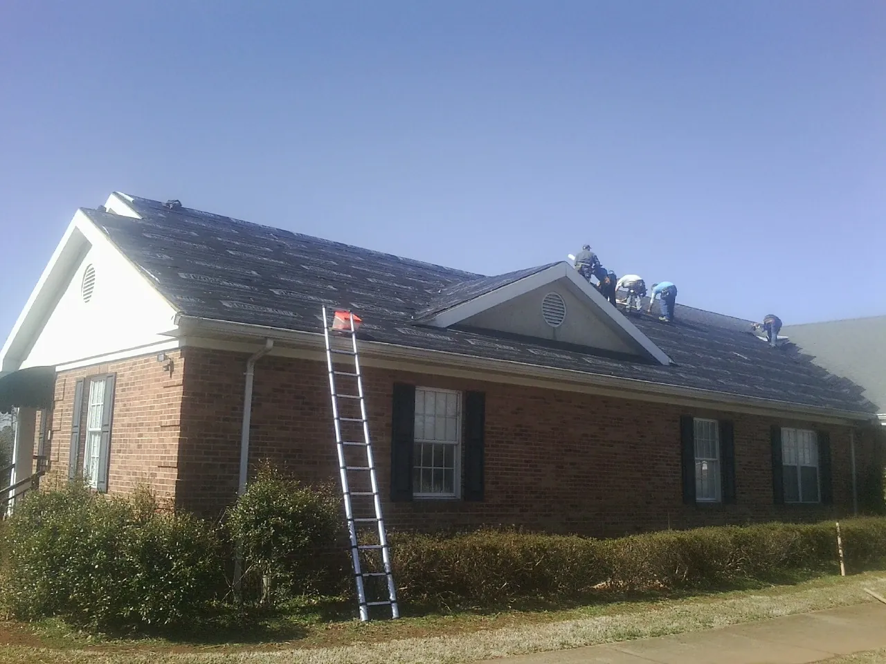 Crew tearing shingles off a brick ranch roof with a ladder set — Metal Roof Repair work by our crew in Bancroft